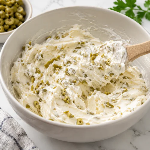 Softened cream cheese mixed with chopped green olives in a bowl on a clean kitchen counter.