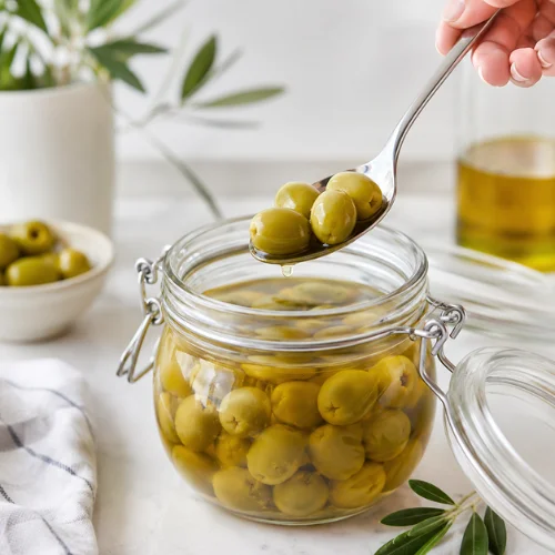 Clean spoon taking green olives from a glass jar filled with brine on a white kitchen counter.